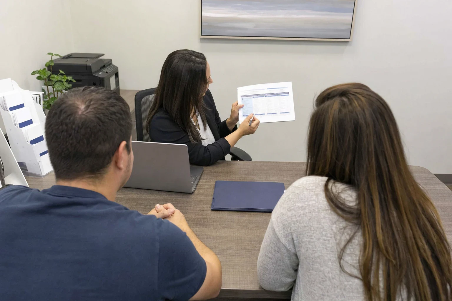 Insurance agent at a desk with a laptop and blue folder shows a printed comparison sheet to a couple seated across from her in a small office with a framed print.