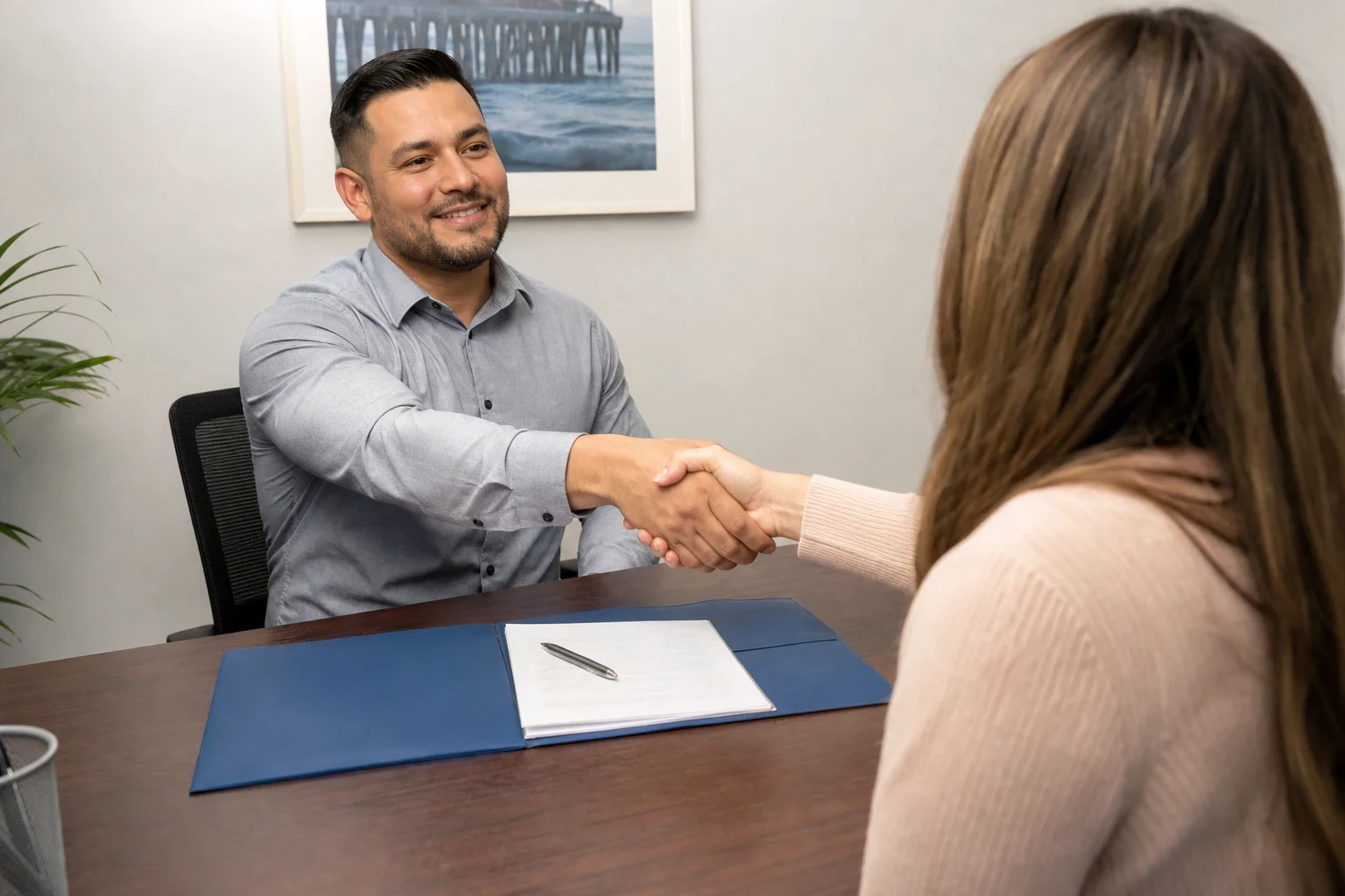 Smiling insurance agent shakes hands with a client across a wooden desk holding a blue folder, papers and pen, with a framed pier photo on the wall behind