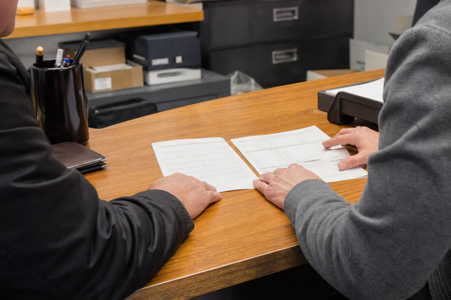 Two people seated at a wooden office desk reviewing two printed documents laid side by side, with a pen cup, wallet and filing cabinets in the background